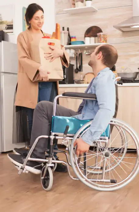 Woman carrying groceries while speaking with a man using a wheelchair in a kitchen.